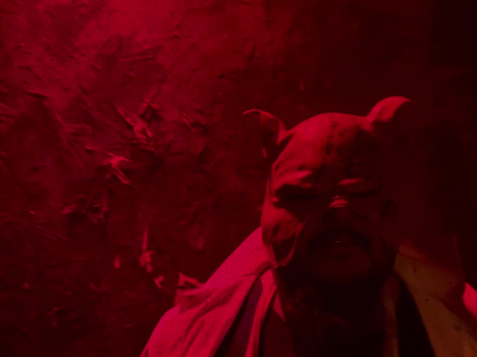 A pig-masked butcher in a stained apron under deep red light at Corn Creepers