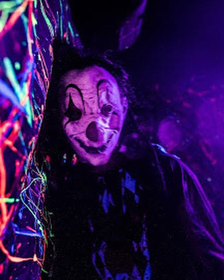 A masked clown with a pale grinning face in a neon-splattered corridor under ultraviolet light