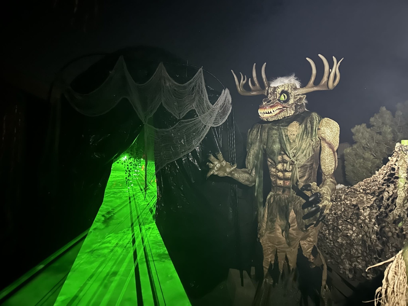 An antlered, fanged creature beside a green-lit, cobwebbed tunnel entrance at Macabre Manor