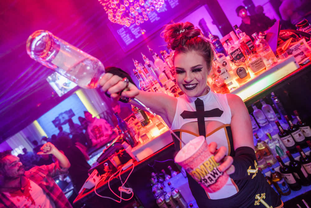 A costumed bartender under pink bar lights pours a bottle into an official Zombie Crawl cup