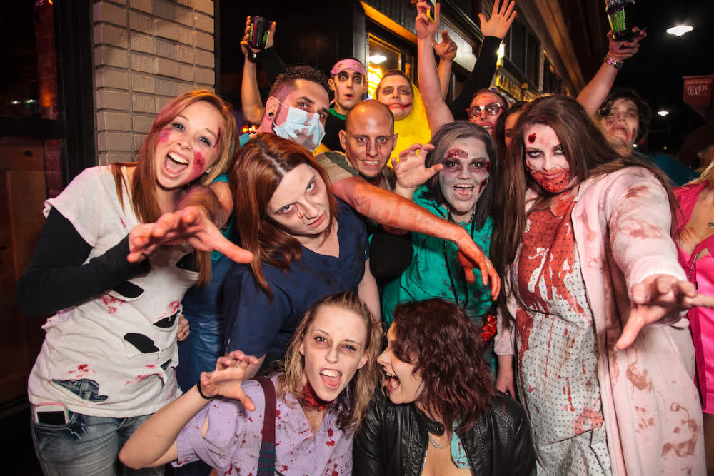 About a dozen adults in bloody zombie makeup crowd together and claw at the camera outside a downtown bar